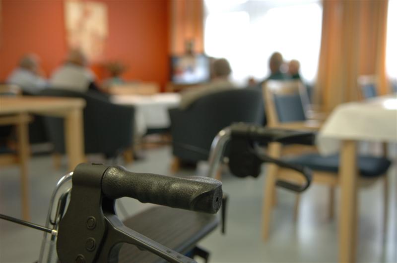 Griffe eines Rollators im Vordergrund, im Hintergrund sitzen Menschen an Tischen